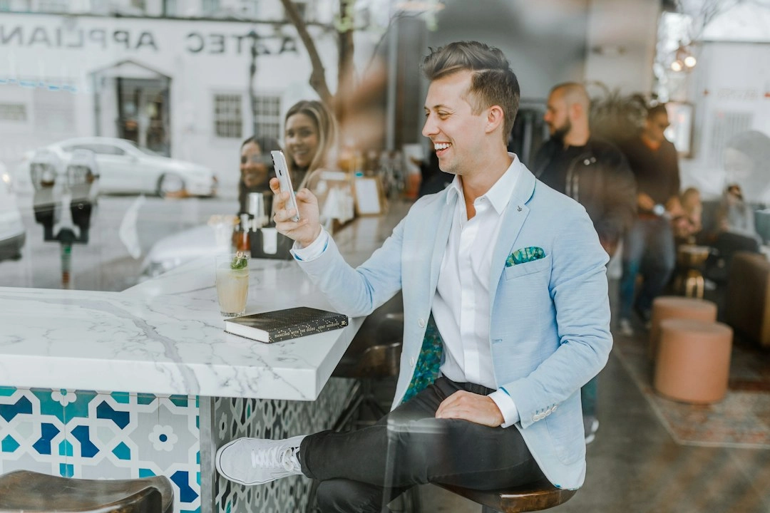 Un uomo con un blazer blu sorride e tiene in mano un segno di pace mentre è seduto al tavolo di un bar visto attraverso una finestra di vetro.